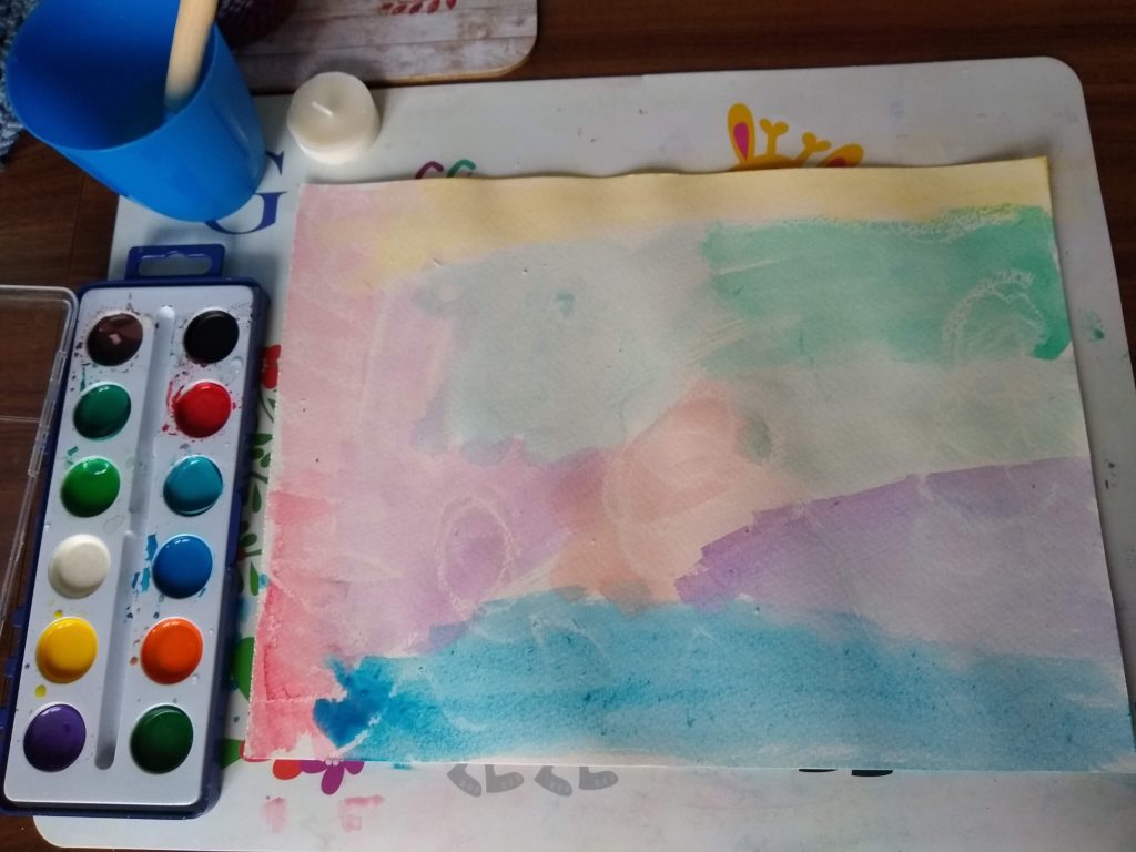 A photo showing water, paints, wax and a picture. The picture is yellow, red, blue, purple, orange and green. It has curls, waves and zigzags.