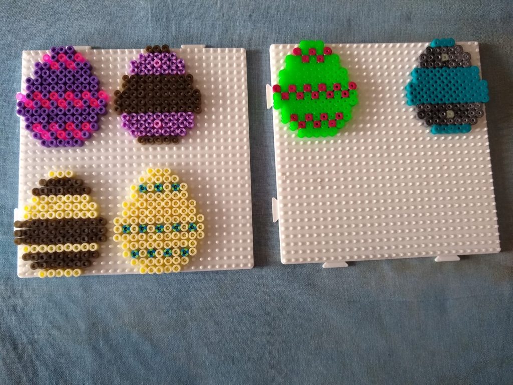 A photo of the Easter eggs before ironing. The first egg is purple with pink zigzags; the second is bronze with pink dots, and purple and pink stripes; the third is cream with bronze stripes, the fourth is cream with blue and green dots, the fifth is green with red zigzags; and the final egg is blue with silver stripes and glow-in-the-dark dots.